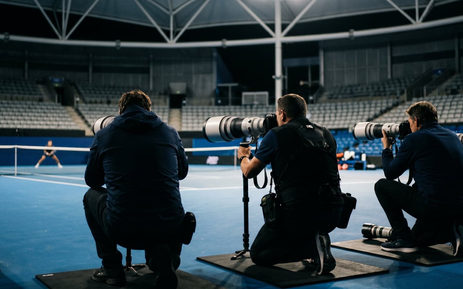 Three professional sports photographers in the courtside photo pit, aiming long white telephoto lenses across an empty cobalt-blue tennis court inside a stadium.
