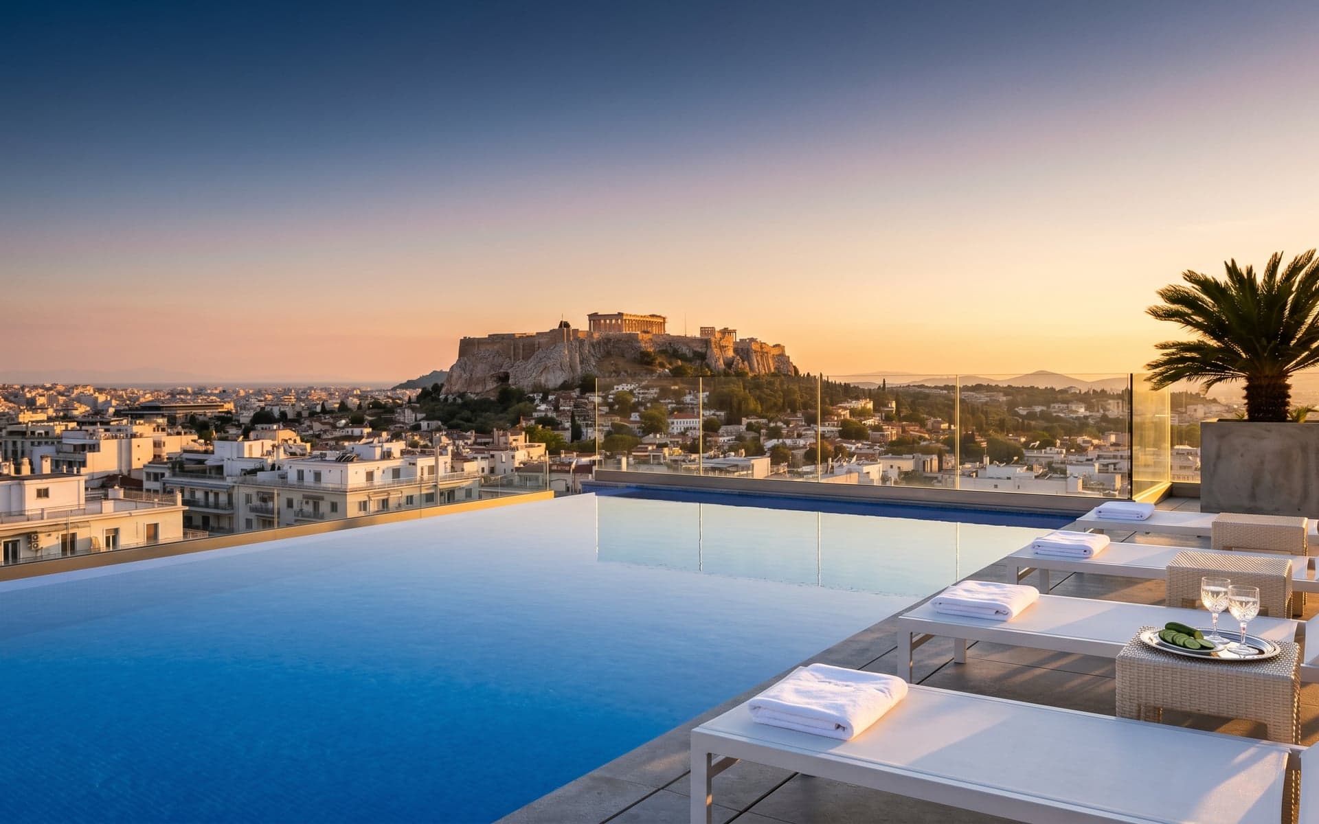 A modern rooftop infinity pool at an Athens 5-star hotel at golden hour, with empty white sun loungers and a postcard view of the Acropolis crowning the Athens skyline.
