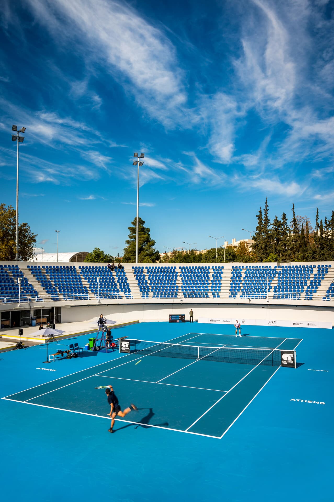 ITC Athens ATP Challenger 2025 — Day 1 match action, player serving on Court 1 with sky and trees behind.